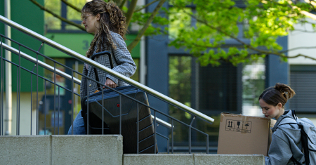 Foto von zwei Personen, die den Eingang zum Wohnheim hochgehen, eine mit Koffer, eine mit einem Karton - Photo of two people walking up towards the entrance to the hall of residence, one carrying a suitcase, the other a box. 