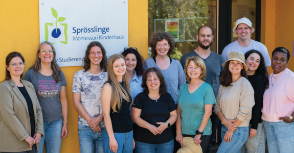 Photo shows the team of Kita Sprösslinge in front of the building and in front of the logo of the kita