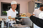 Foto von einem Koch, der das Essen an einen Studierenden (nur Arm zu sehen) reicht, das Foto ist aus dem Food-Truck