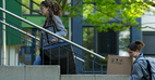 Foto von zwei Personen, die den Eingang zum Wohnheim hochgehen, eine mit Koffer, eine mit einem Karton - Photo of two people walking up towards the entrance to the hall of residence, one carrying a suitcase, the other a box. 